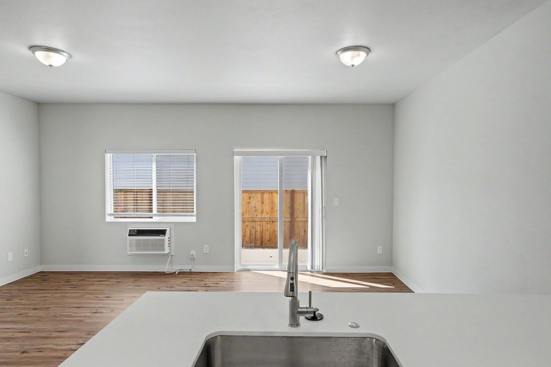 Empty modern room with gray walls, wood floor, window A/C unit, and sliding glass door by a kitchen sink.