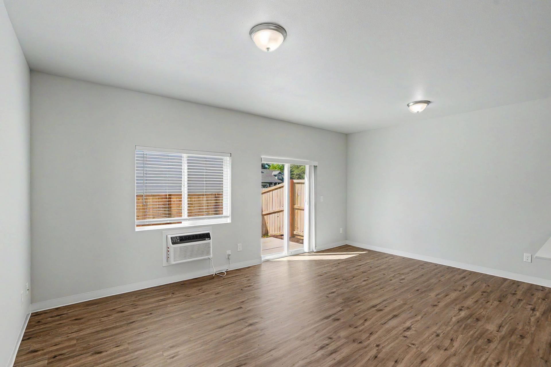 Empty white living room with wood floors, two windows, a sliding glass door, and ceiling lights