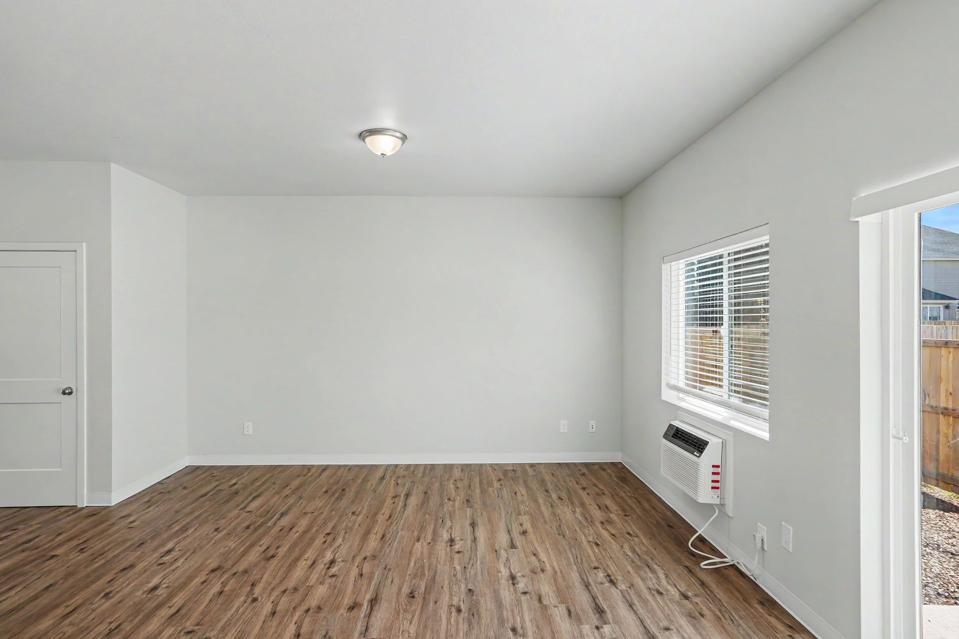 Empty white living room with wood floors, window blinds, and a glass patio door