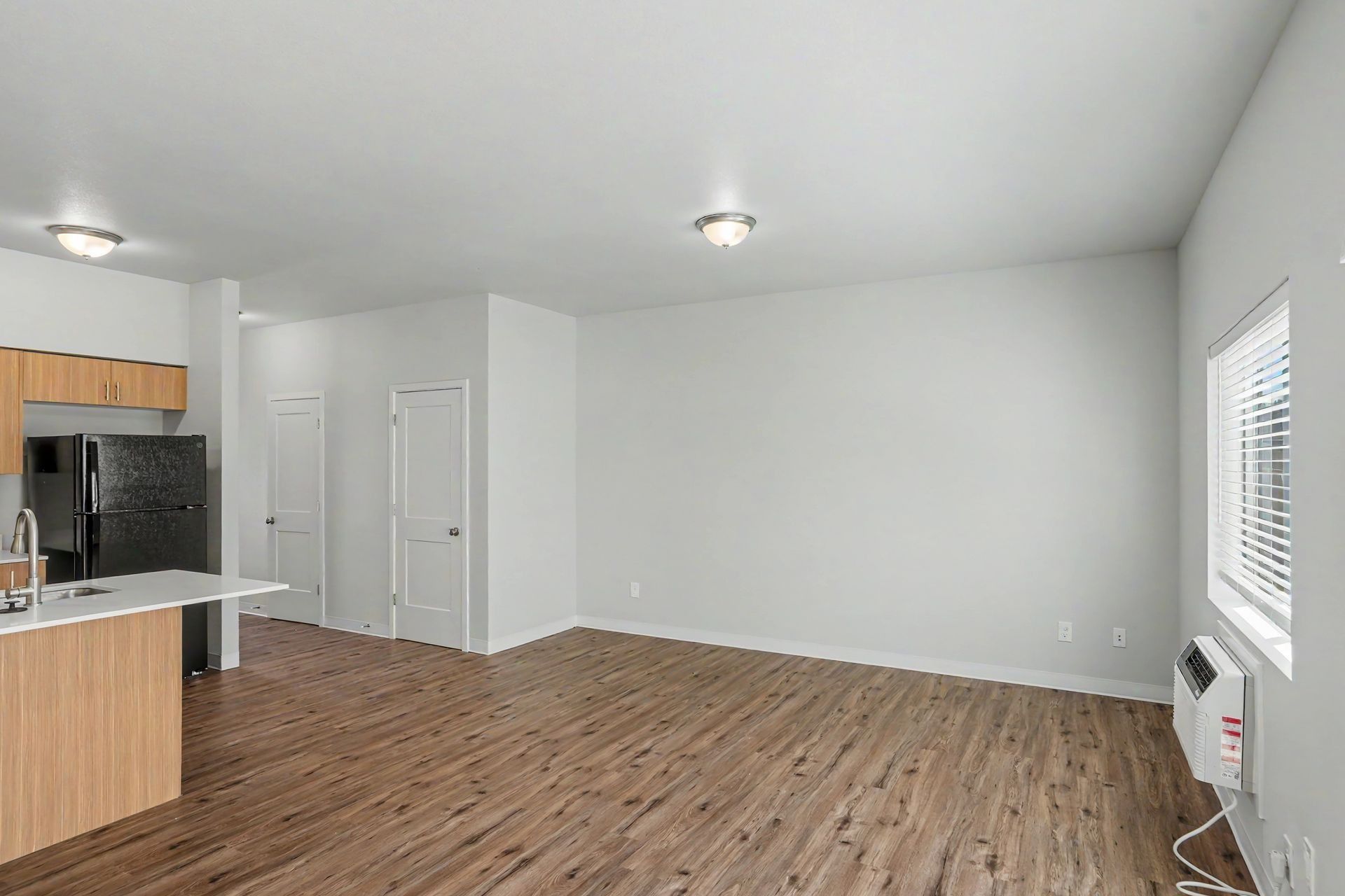 Empty open-plan living room with wood floors, white walls, kitchen area, and a window air conditioner.