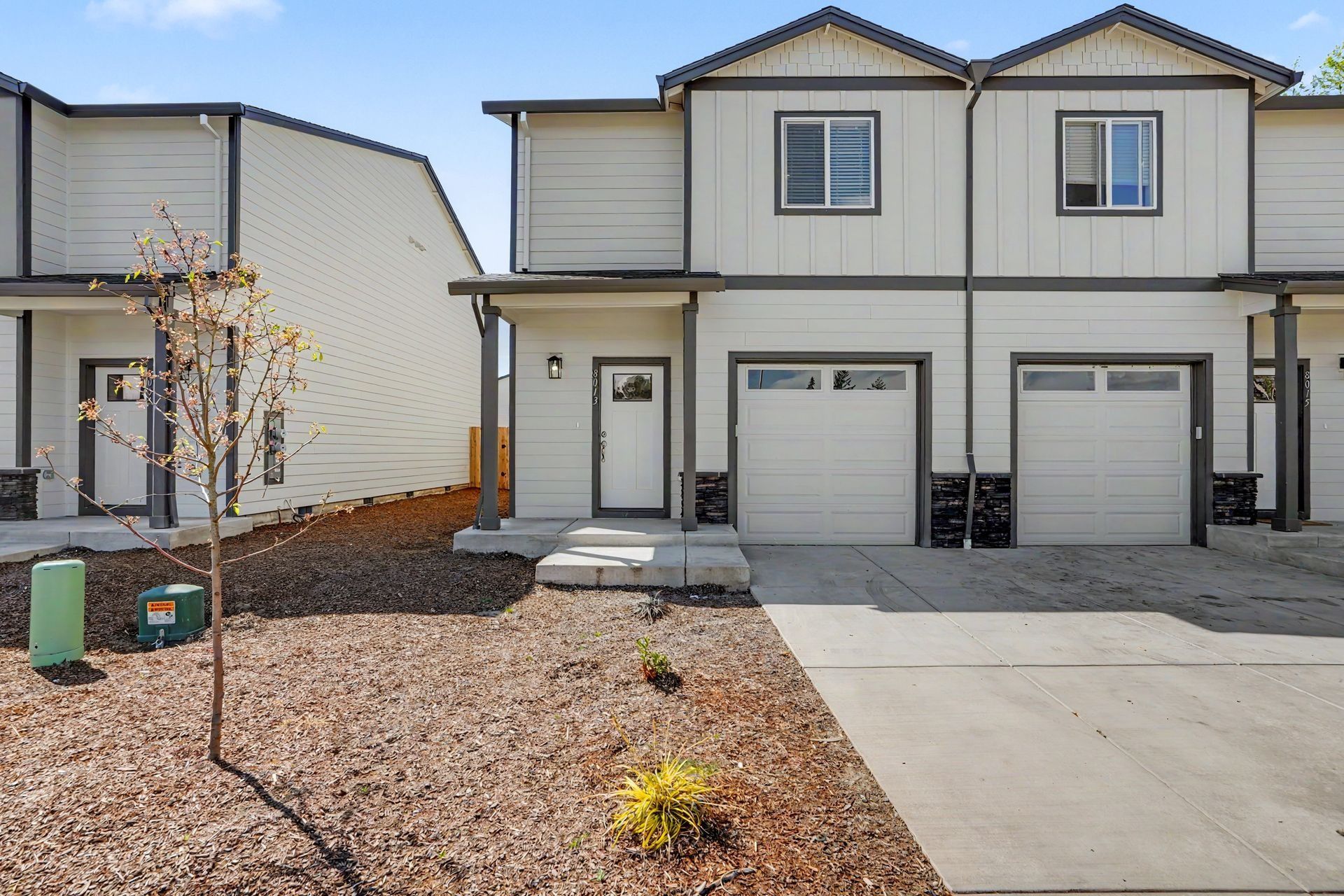 Two-story beige townhouse with two garages, a small front porch, and a gravel yard in a suburban neighborhood