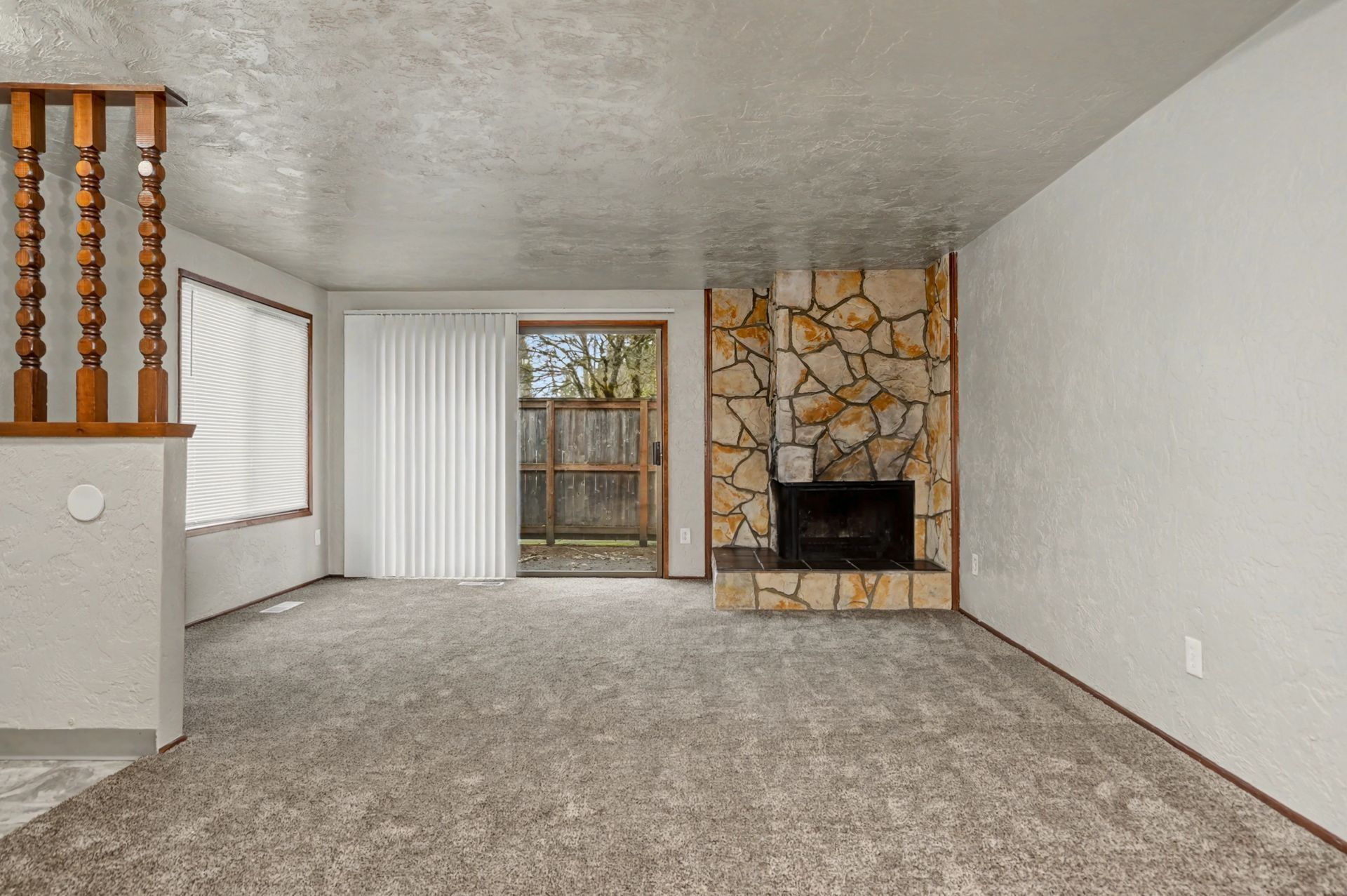 Empty living room with carpet, stone fireplace, and large windows with vertical blinds