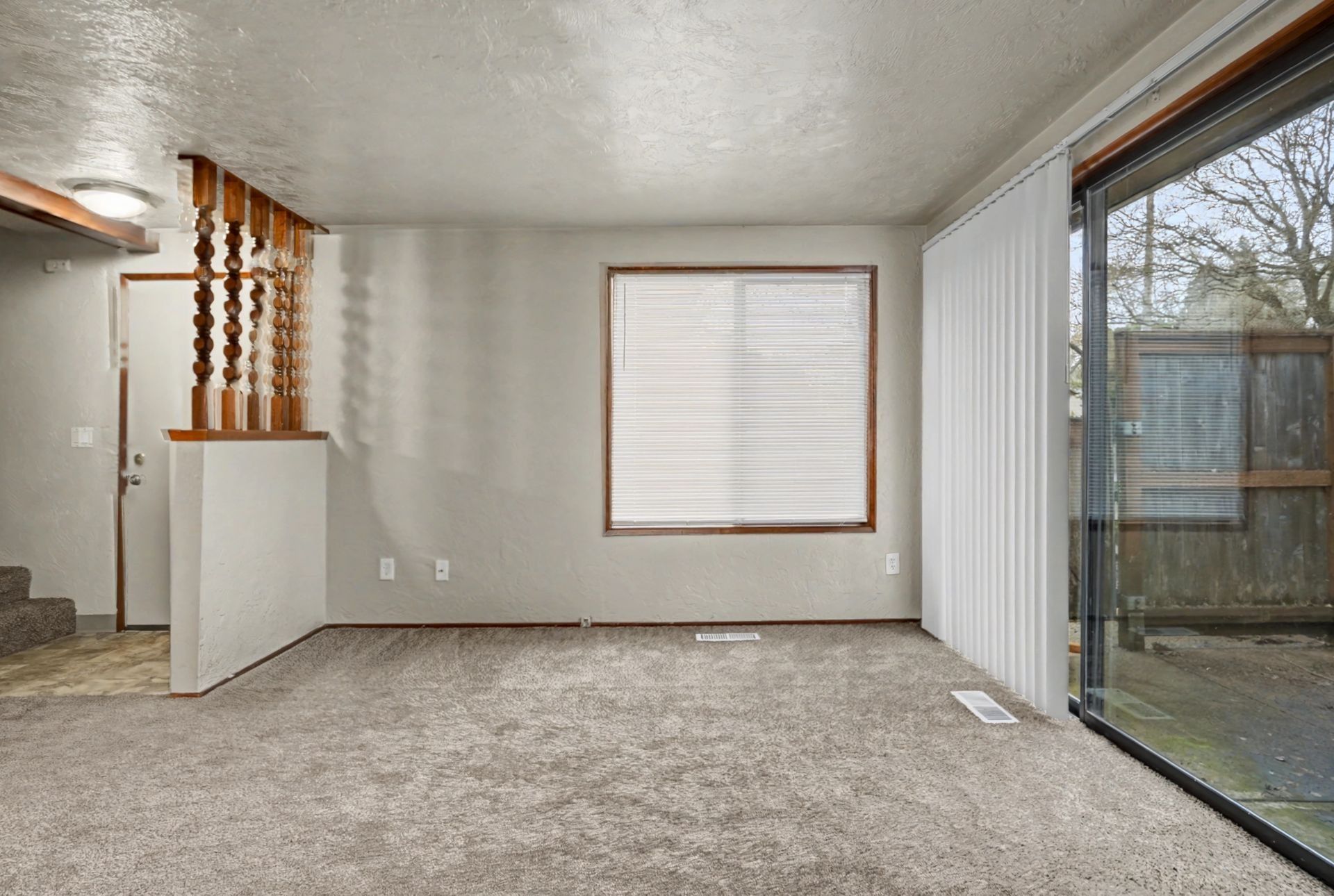 Empty carpeted living room with sliding glass door, window blinds, and staircase railing on the left