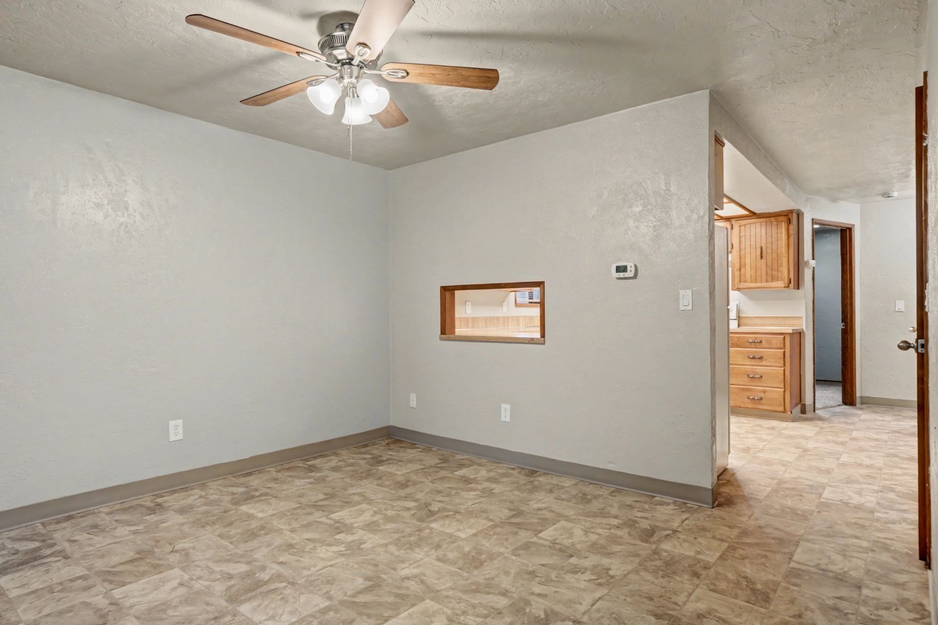 Empty gray bedroom with carpet, ceiling fan, small window, and doorway to a kitchen area