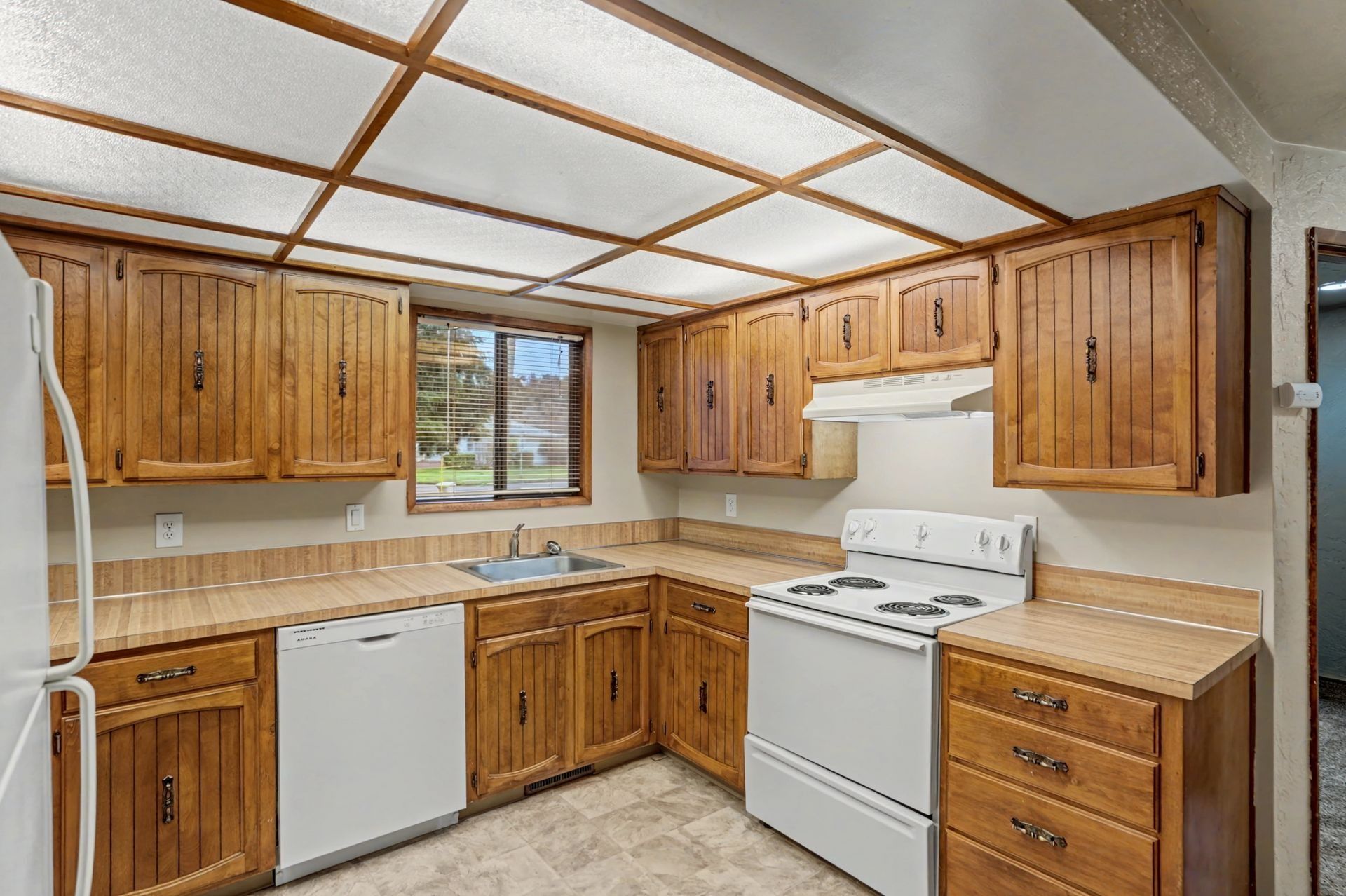 Wooden kitchen with white appliances, beige countertops, and a window over the sink
