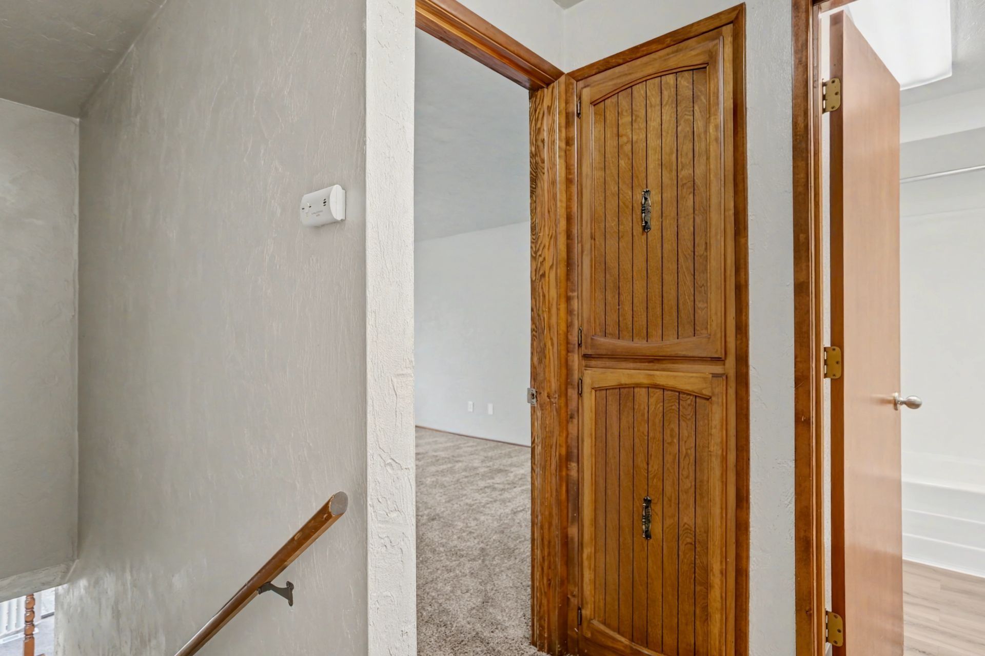 Hallway with open wooden door, stair rail, and light-colored walls leading into a bedroom.