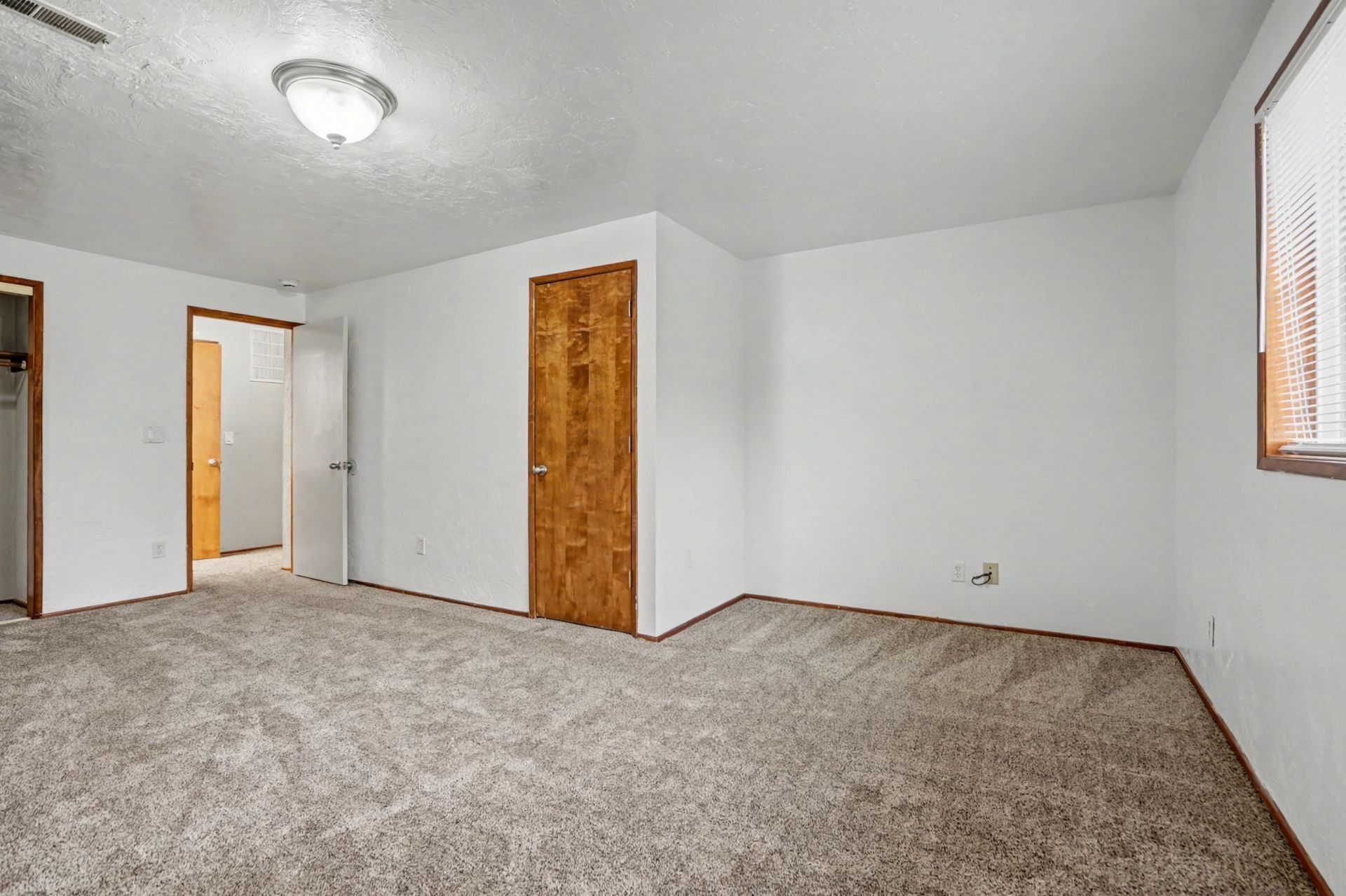 Empty carpeted room with white walls, two wooden doors, and a window with blinds.
