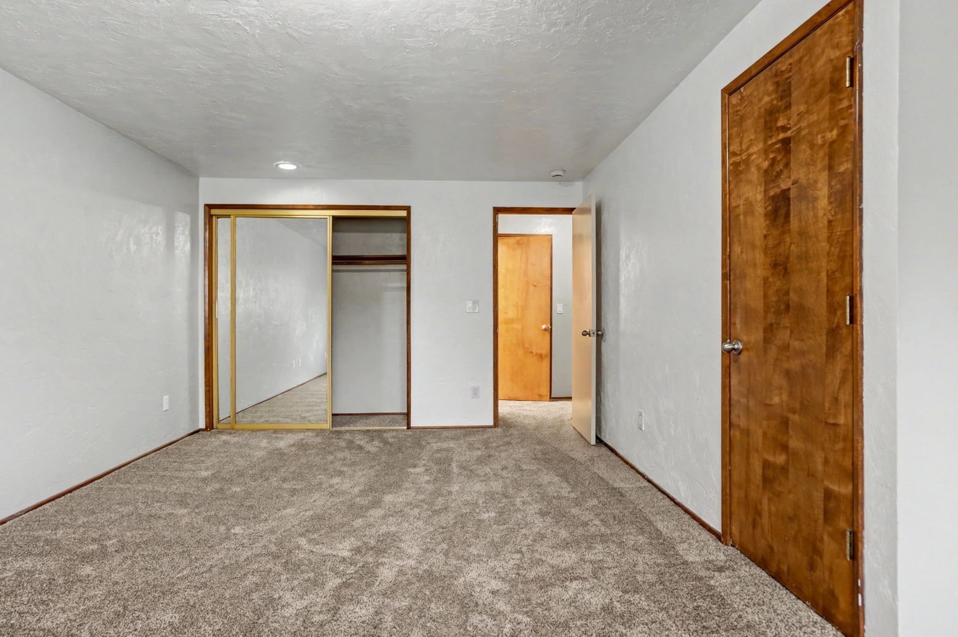 Empty carpeted bedroom with white walls, mirrored closet doors, and a wood door open to a hallway