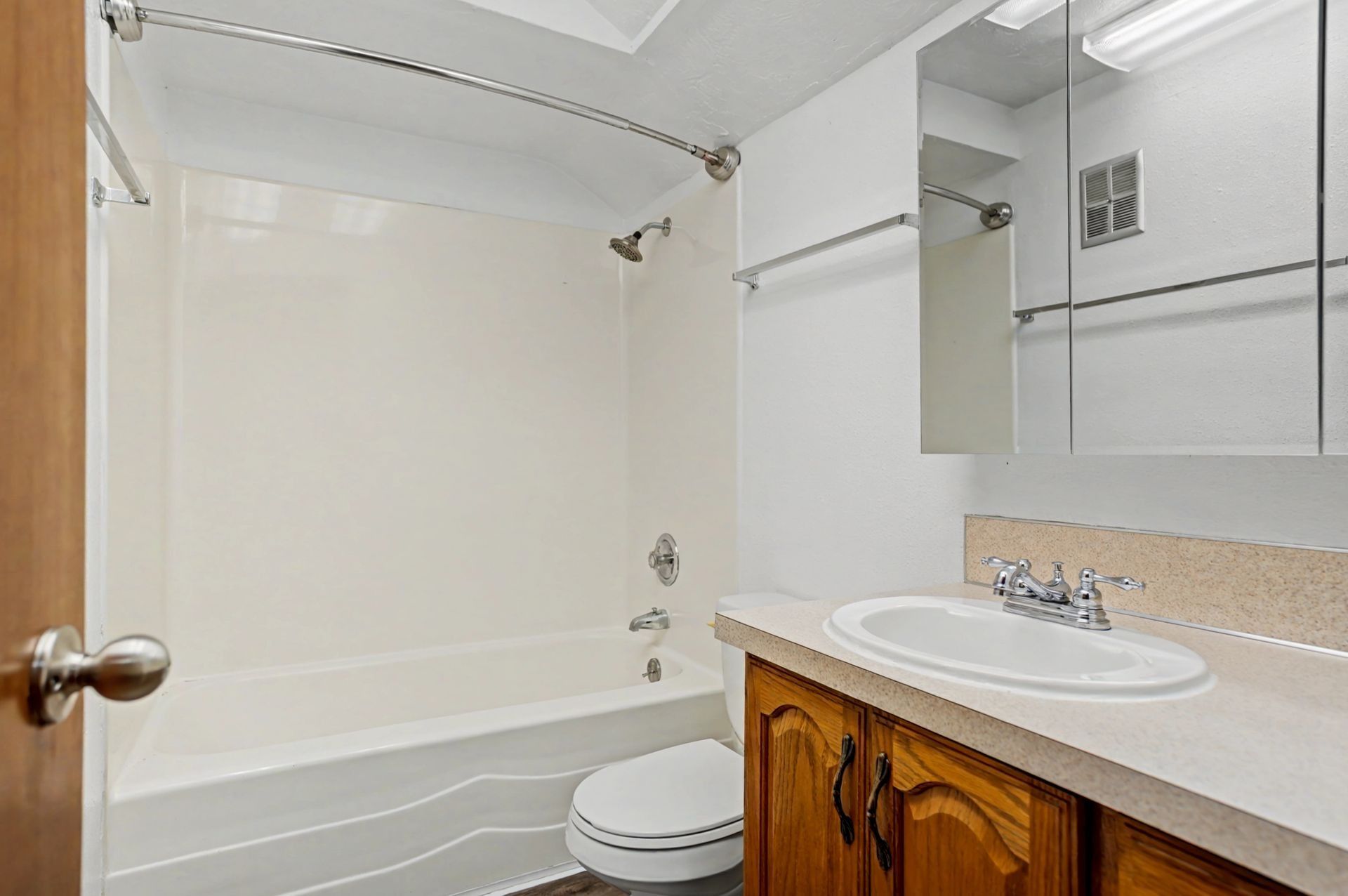 Small bathroom with white tub and toilet, wood vanity, mirror, and beige tile countertop.