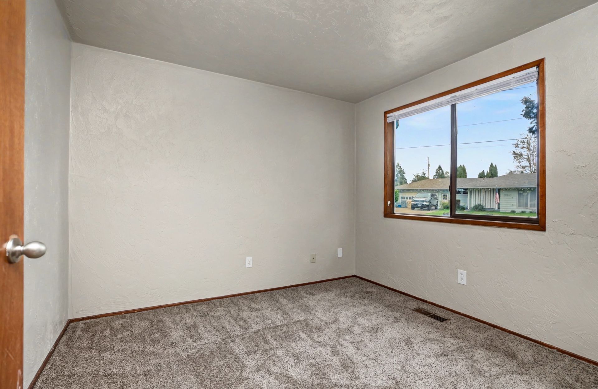 Empty carpeted bedroom with white walls, a wood-framed window, and a closed door.