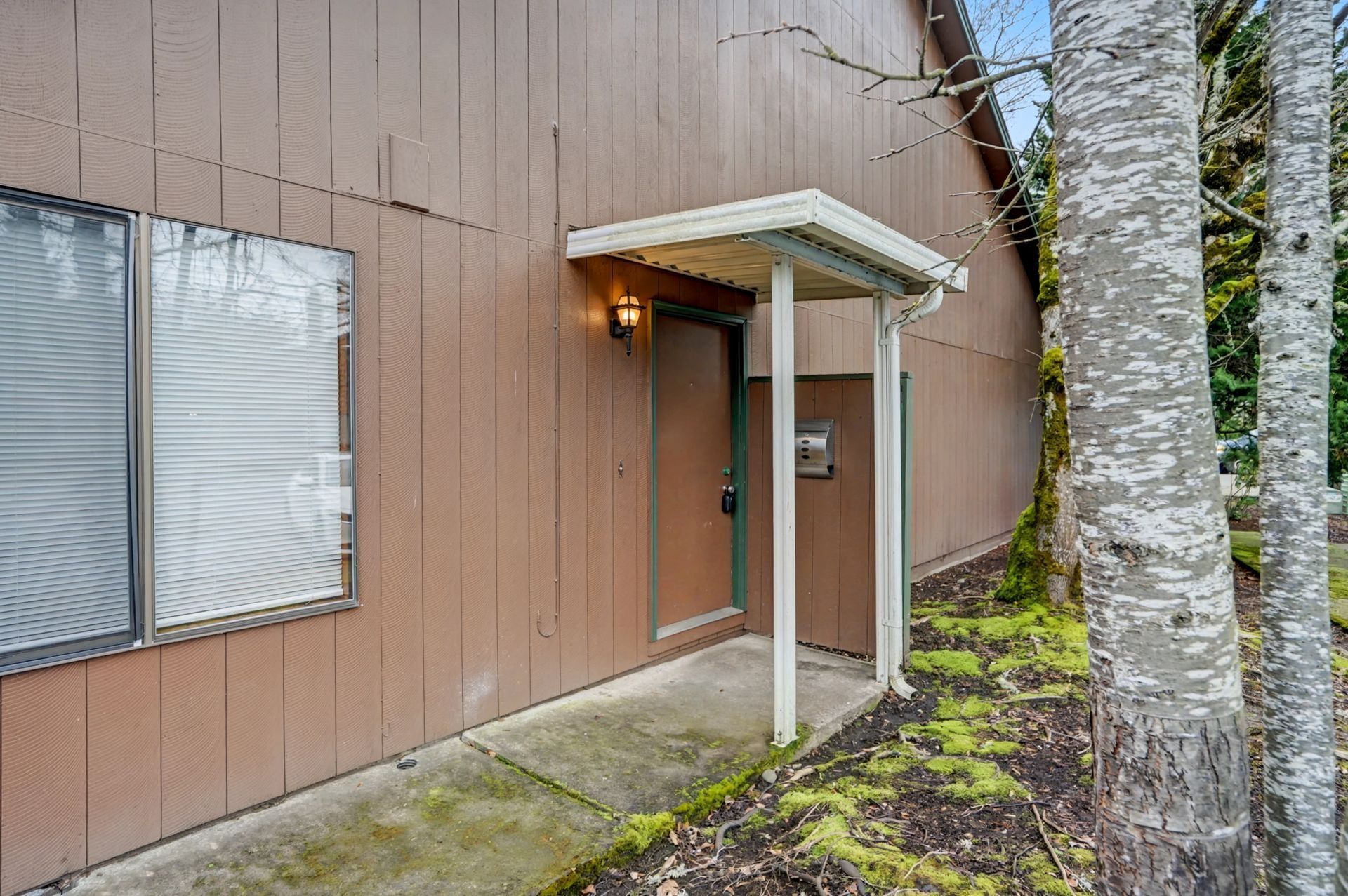 Exterior of a brown building entrance with a small awning, lit doorway, and trees nearby
