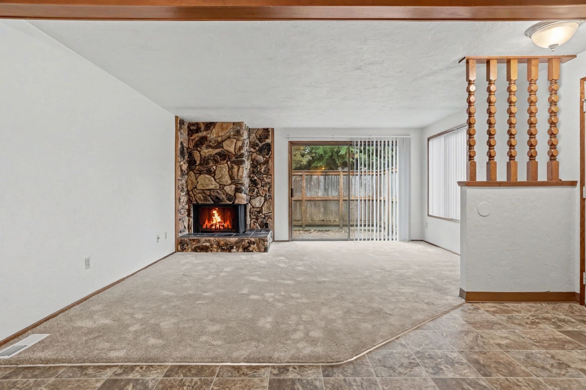 Empty living room with stone fireplace, beige carpet, sliding glass door, and wood stair railing
