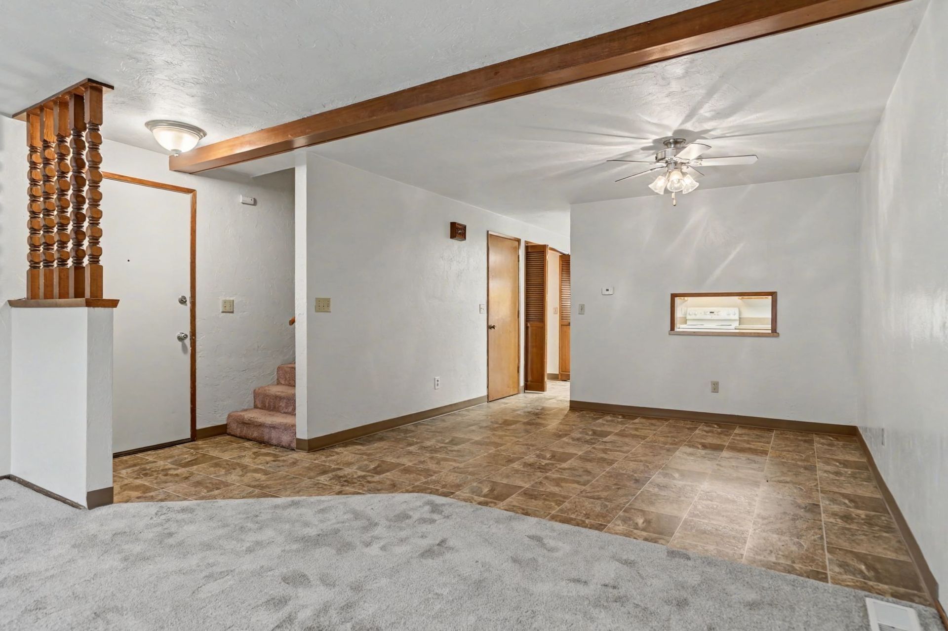 Empty living room with white walls, stone tile floor, exposed wood beam, and staircase to the left