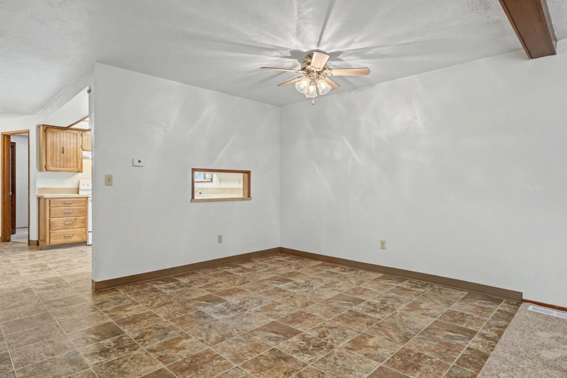 Empty room with white walls, tiled floor, ceiling fan, and a small pass-through window to a kitchen area