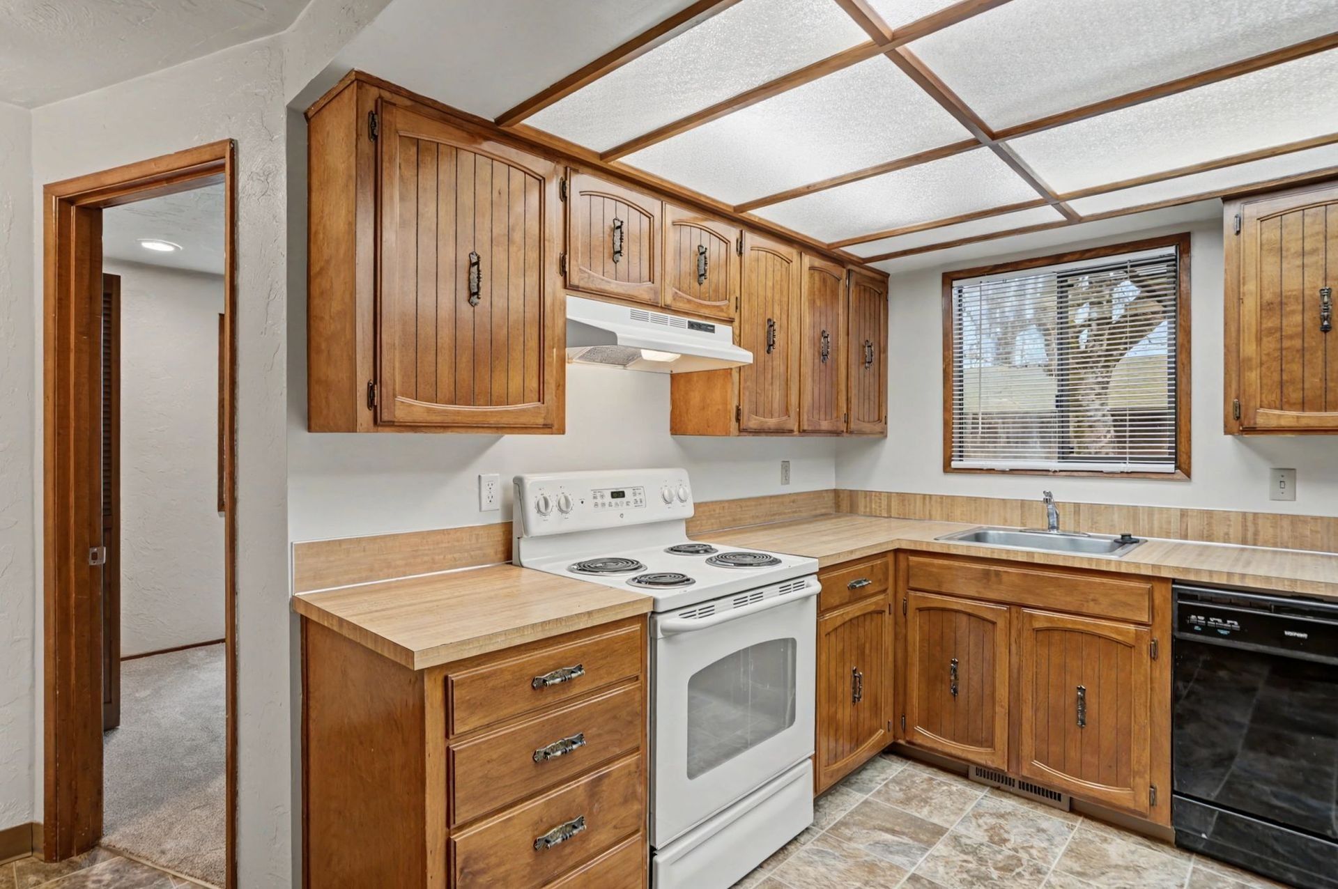 Small kitchen with wood cabinets, white stove, black dishwasher, and beige tile floor.