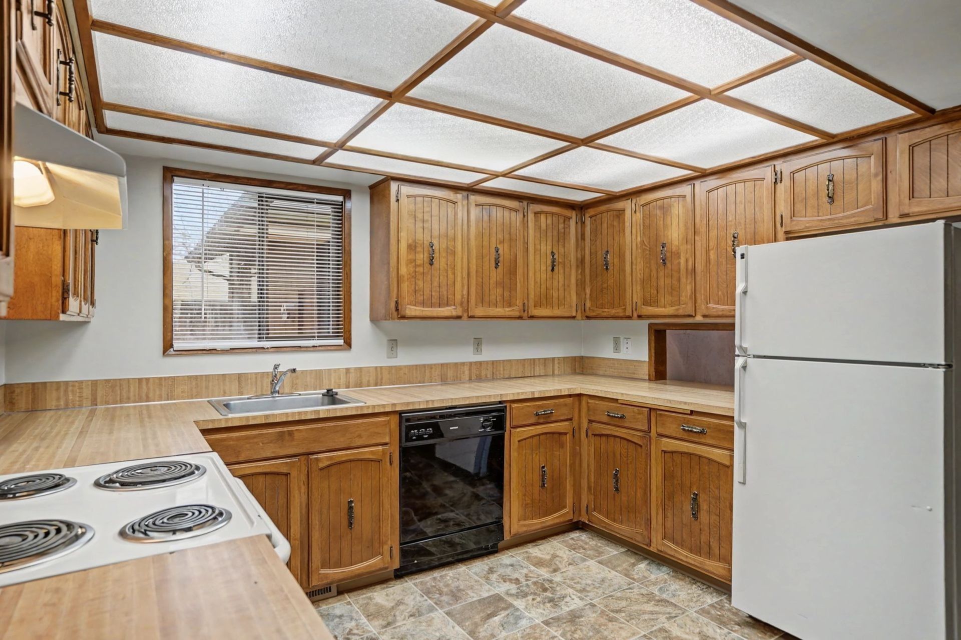 Wooden kitchen with white appliances, tiled floor, and large window under a grid ceiling