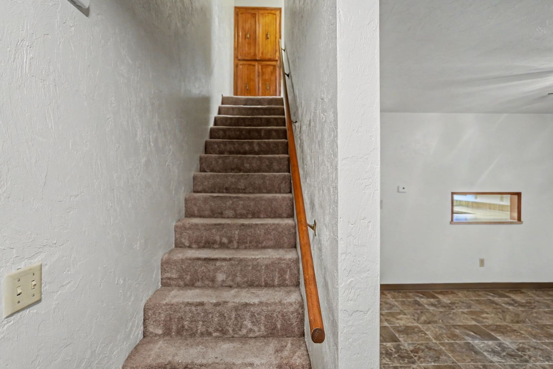 Carpeted staircase with wooden handrail leading to a closed wooden door in a white hallway
