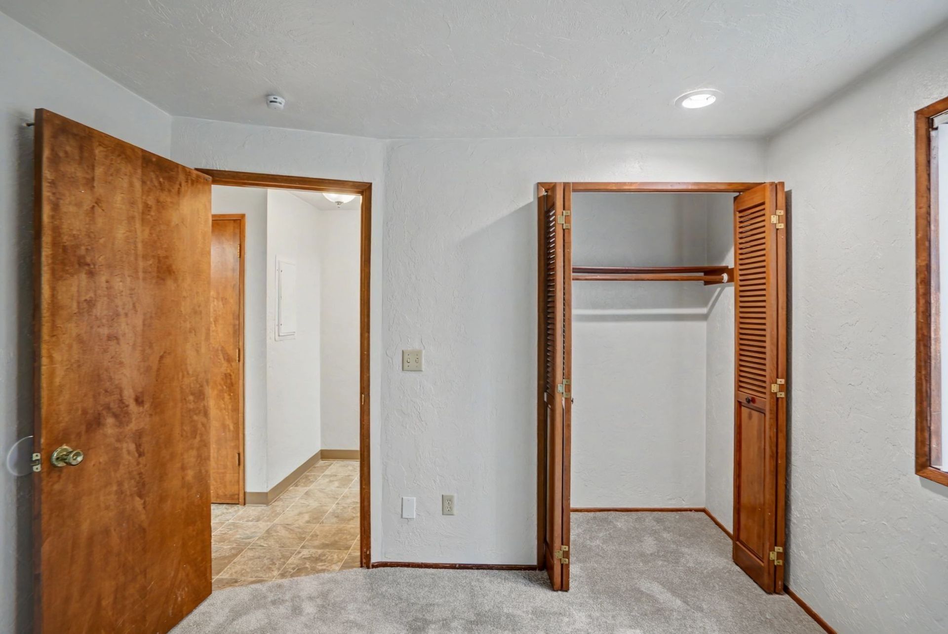 Empty carpeted bedroom with open wooden door and open closet, white walls and ceiling lights