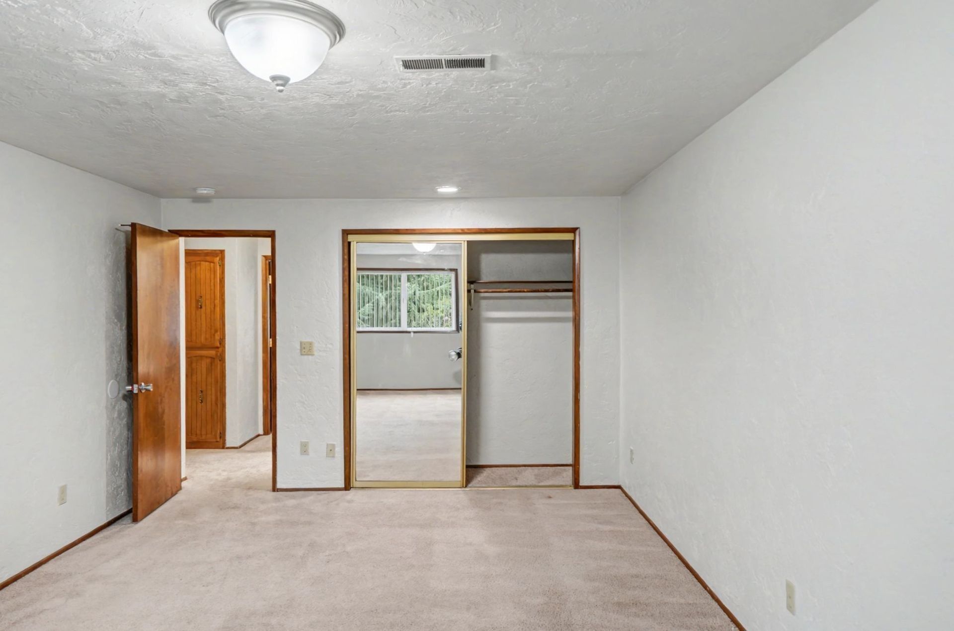 Empty beige bedroom with open closet, doorways, and a window on the far wall.