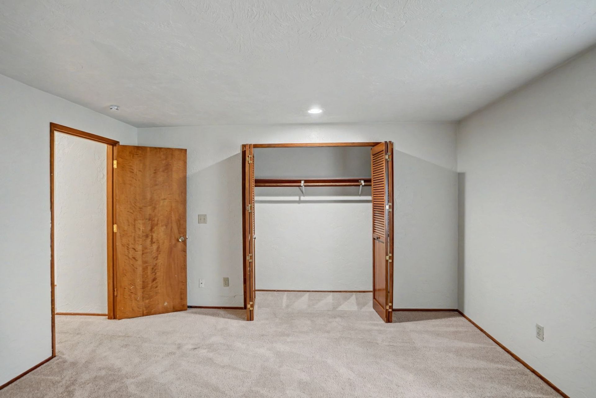 Empty beige bedroom with open closet and wooden door, carpeted floor, and white walls