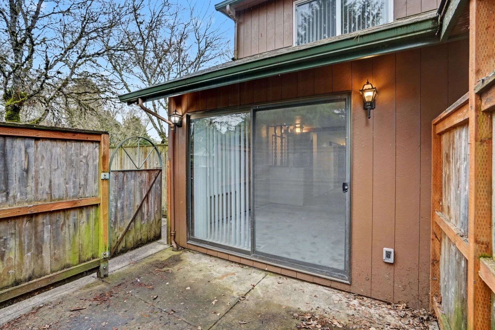 Backyard patio with sliding glass doors, wooden fence, and an overhead deck lit by a wall lantern