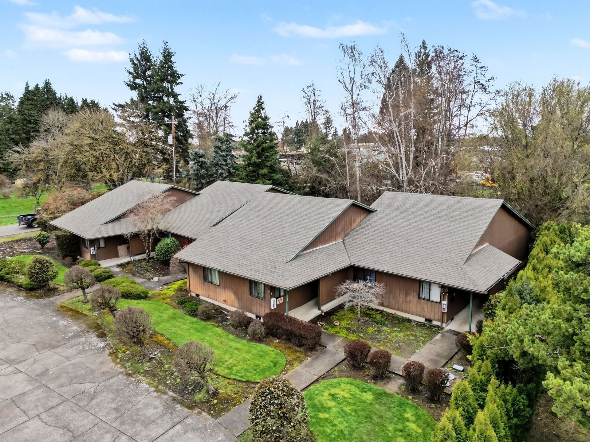 Three small brown houses with gray roofs beside a paved driveway, surrounded by green trees and shrubs