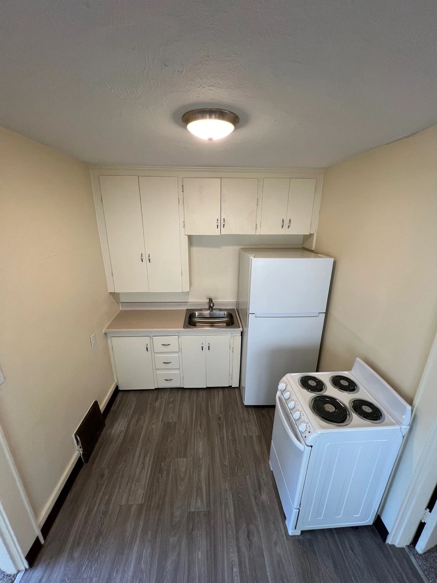 Small kitchen with white cabinets, refrigerator, stove, sink, and gray flooring under a ceiling light.