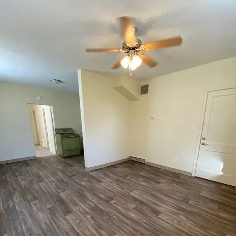 Empty room with wood-look flooring, white walls, ceiling fan, and doorway to a kitchen area