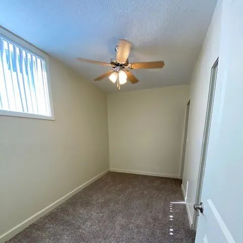 Empty narrow room with beige walls, carpet, ceiling fan, and a small window.