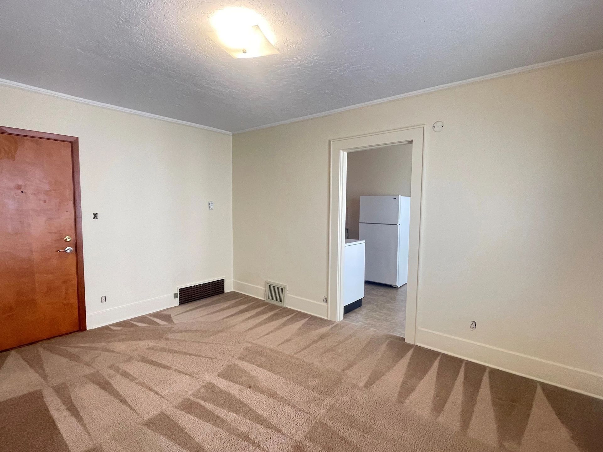 Empty beige room with carpet, wooden door, and open doorway to a kitchen area