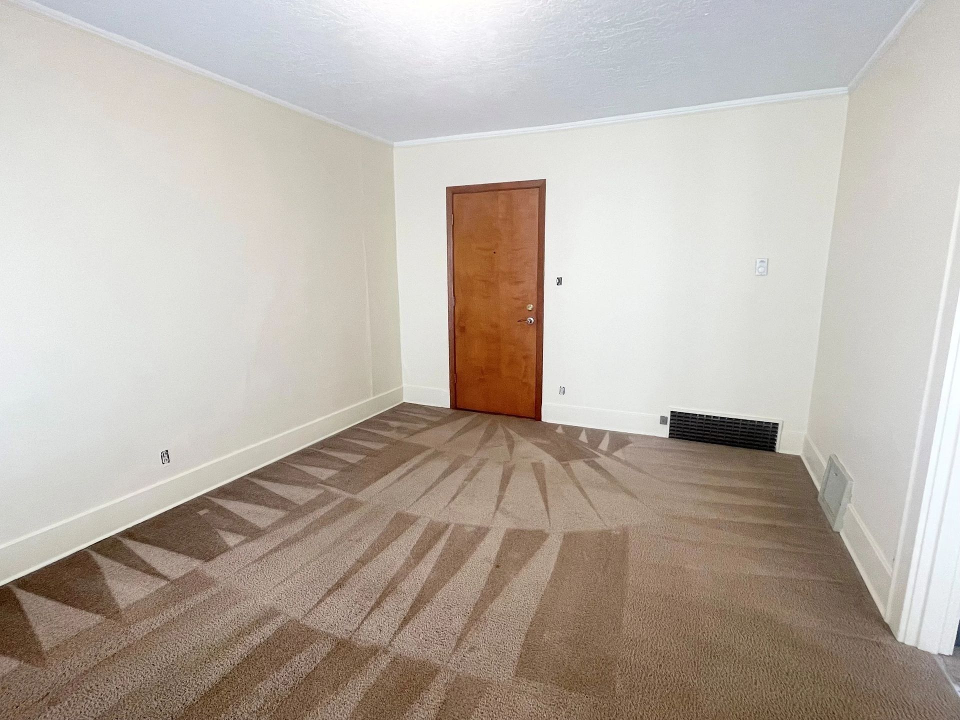 Empty beige room with carpet, white walls, wooden door, and a small vent near the corner