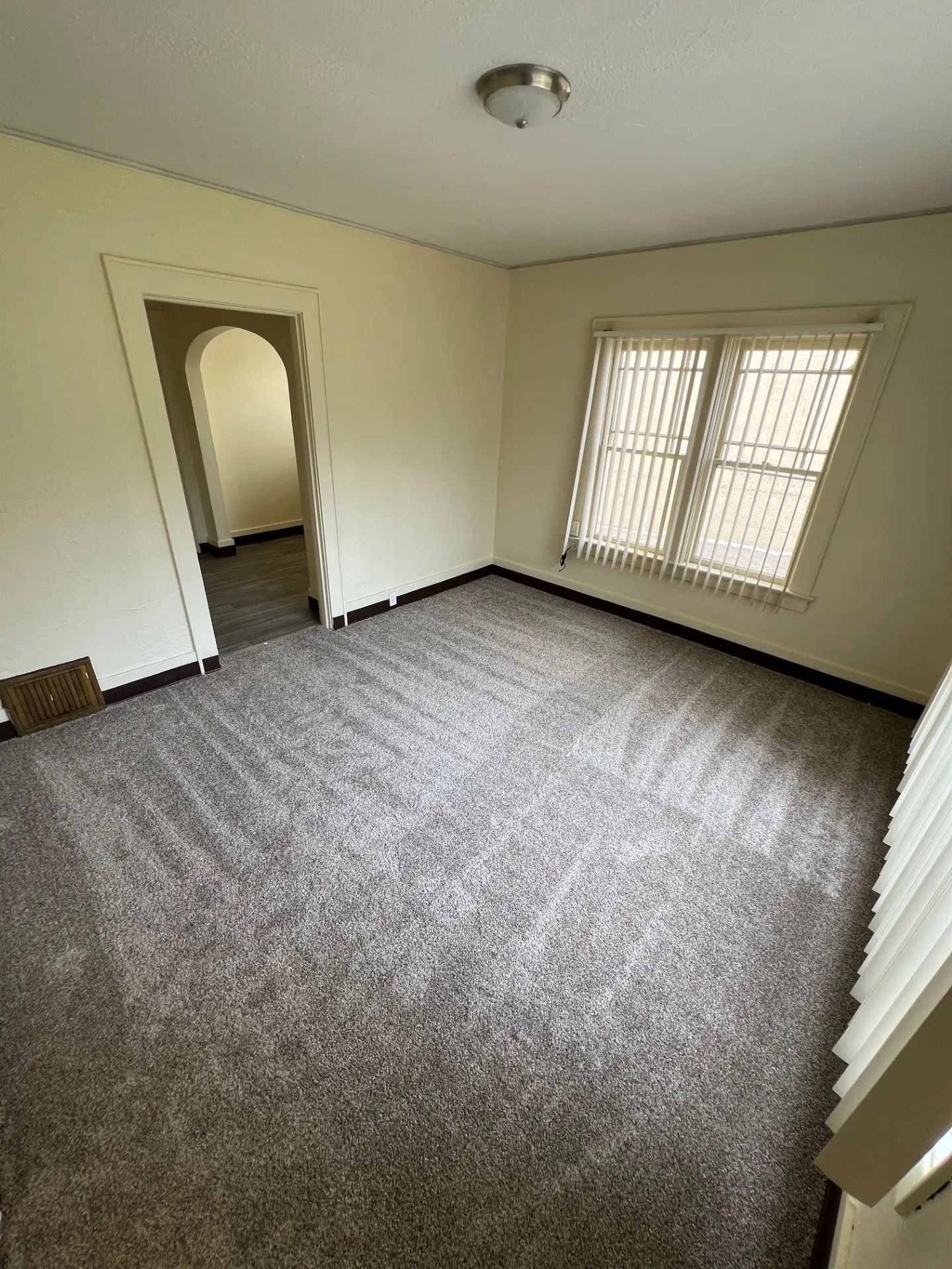 Empty carpeted room with beige walls, arched doorway, and window blinds letting in light.