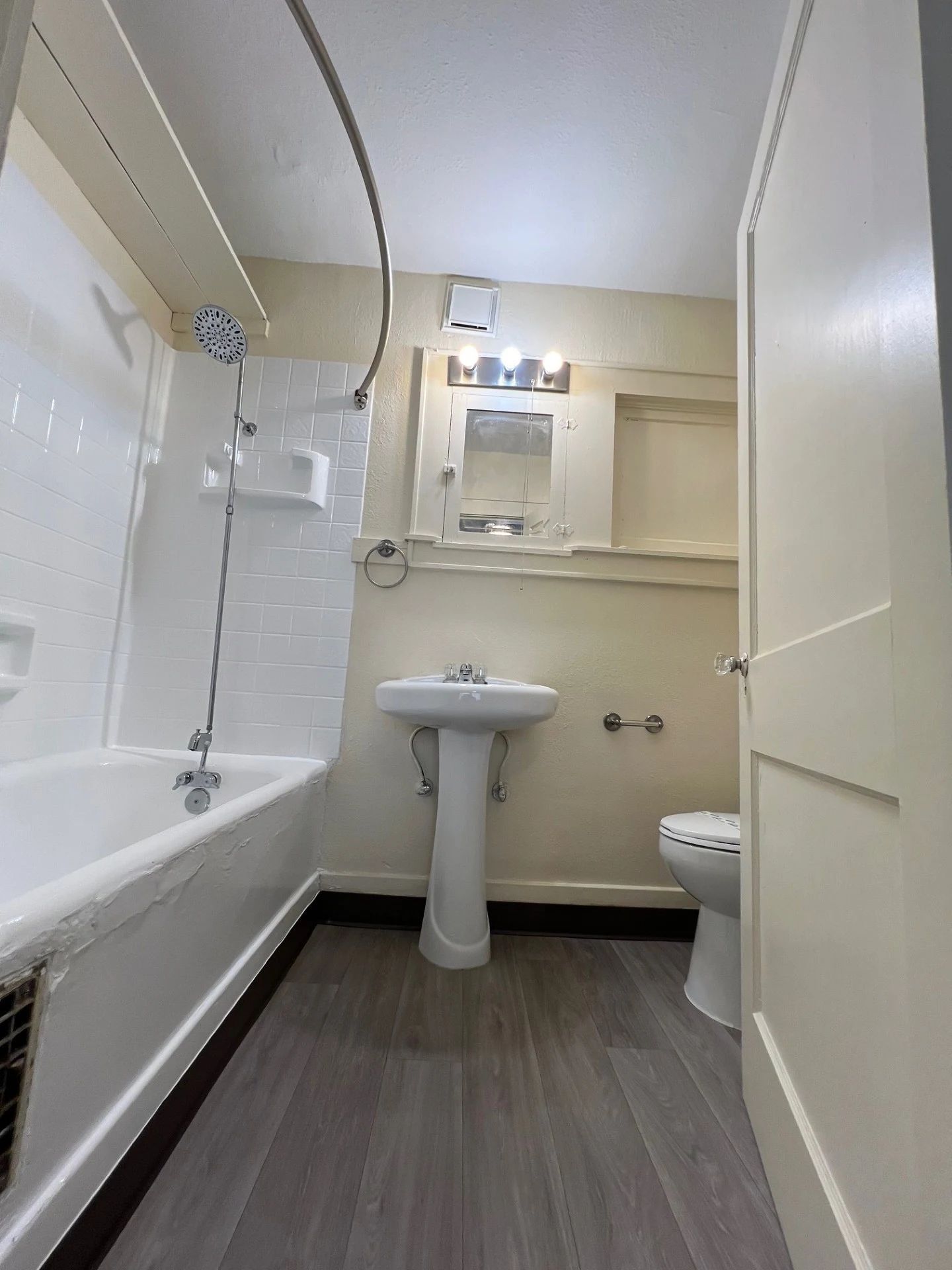 Small bathroom with white tub, pedestal sink, mirror cabinet, and toilet on wood-look flooring.