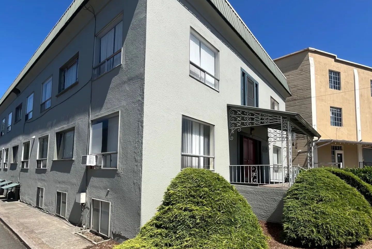 Gray apartment building with white-trimmed windows and a small entry awning on a sunny street corner