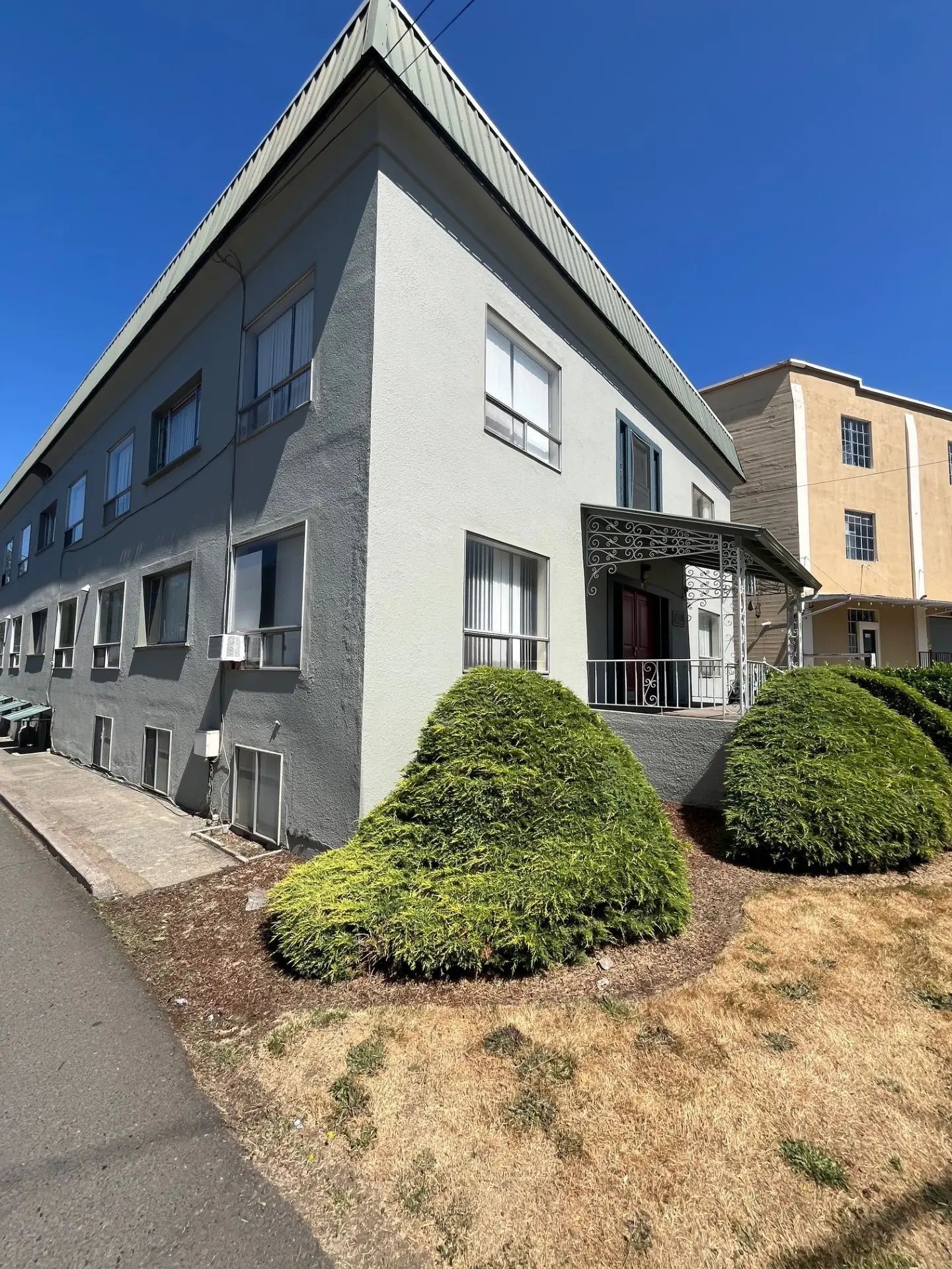 Gray apartment building with black roof beside a sidewalk and dry grass, with shrubs near the entrance