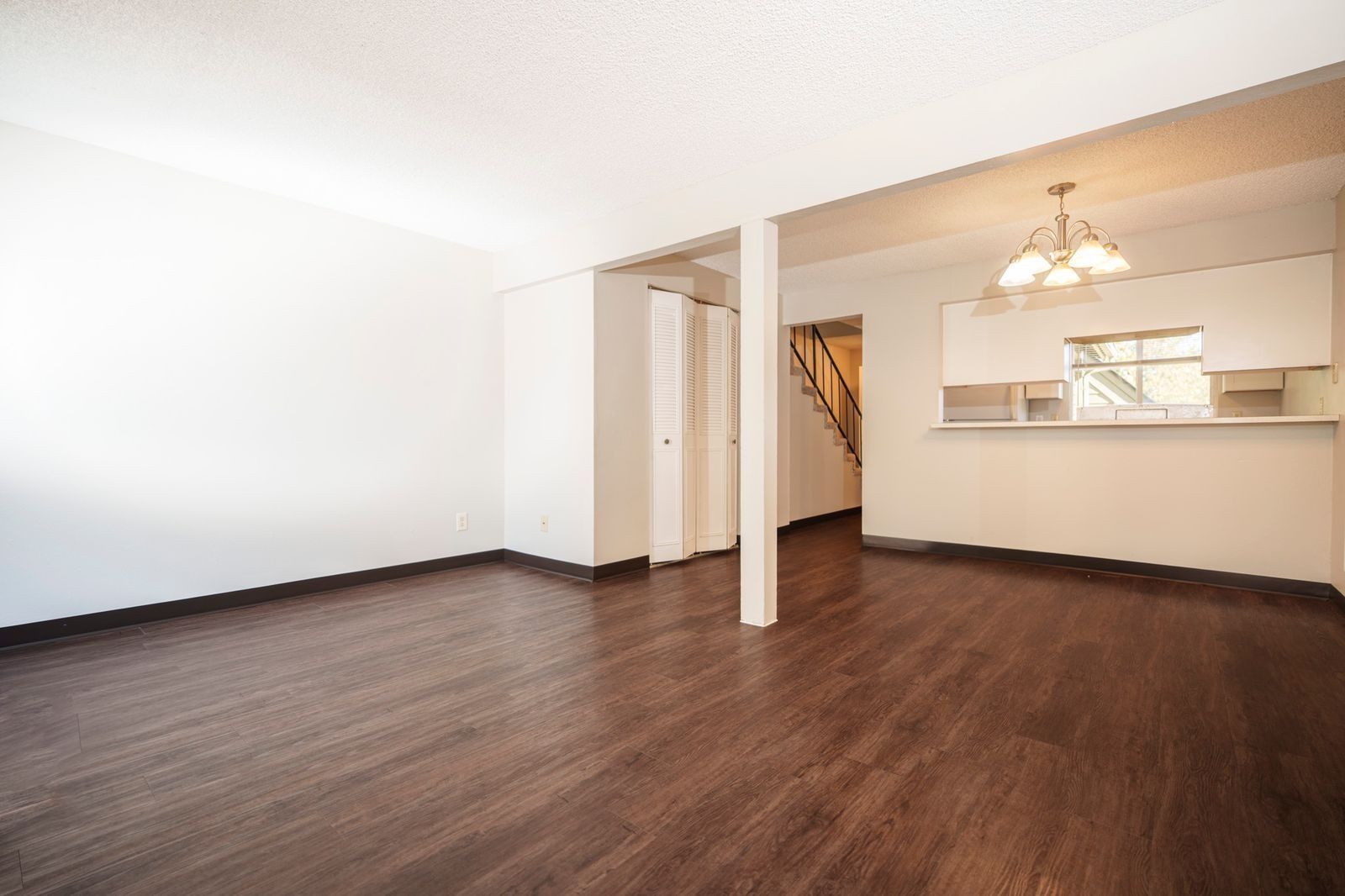 Empty living room with dark wood floors, white walls, and a pass-through opening to a kitchen area.