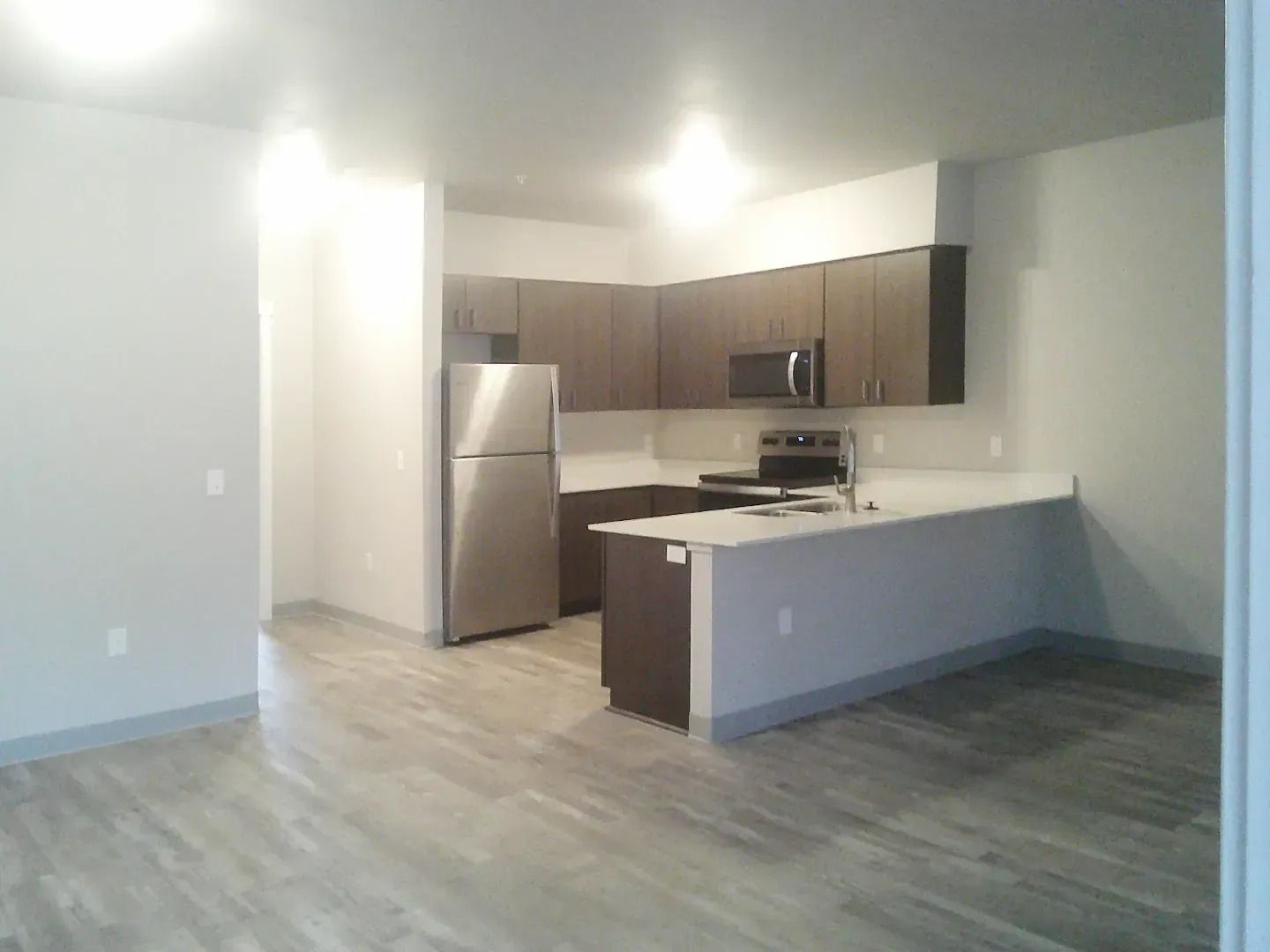 Empty modern kitchen and living area with gray wood floors, white counters, and dark cabinets