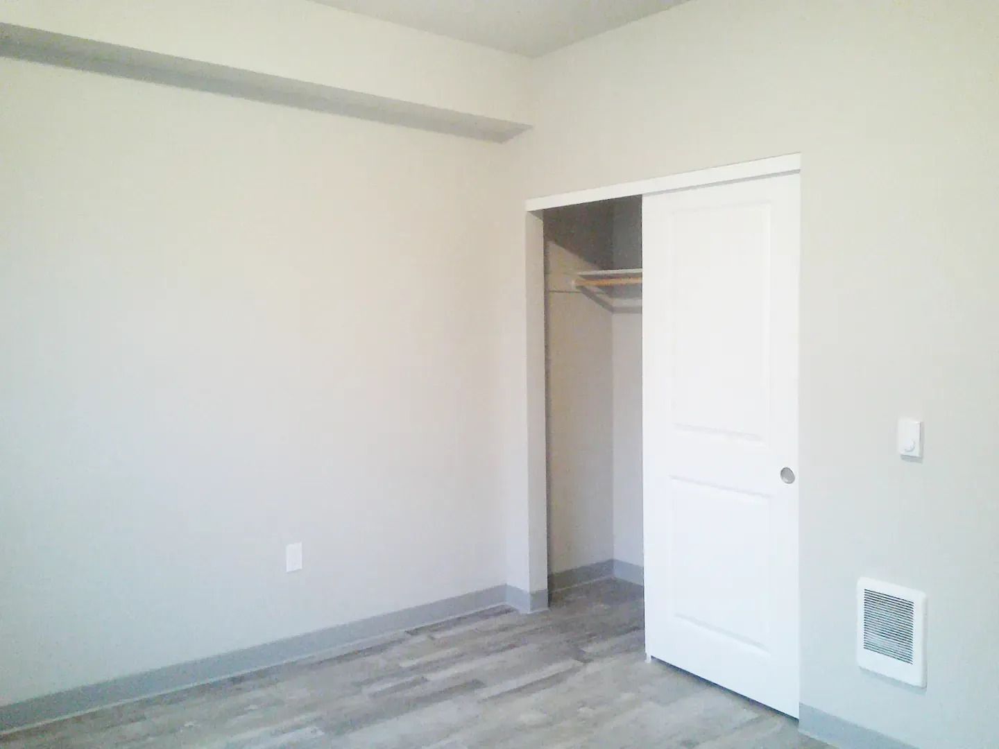 Empty white bedroom with open closet and sliding door, gray wood-look floor, and a small wall vent