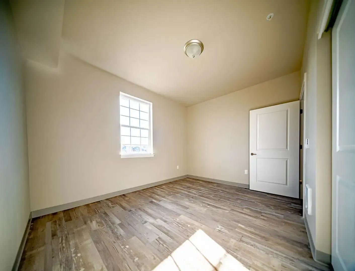 Empty beige bedroom with wood floors, a small window, and a closed white door