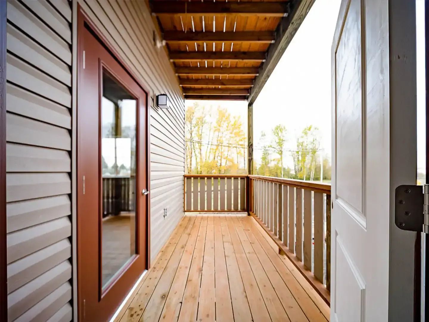 Covered wooden porch with railing, door, and windows beside a house.