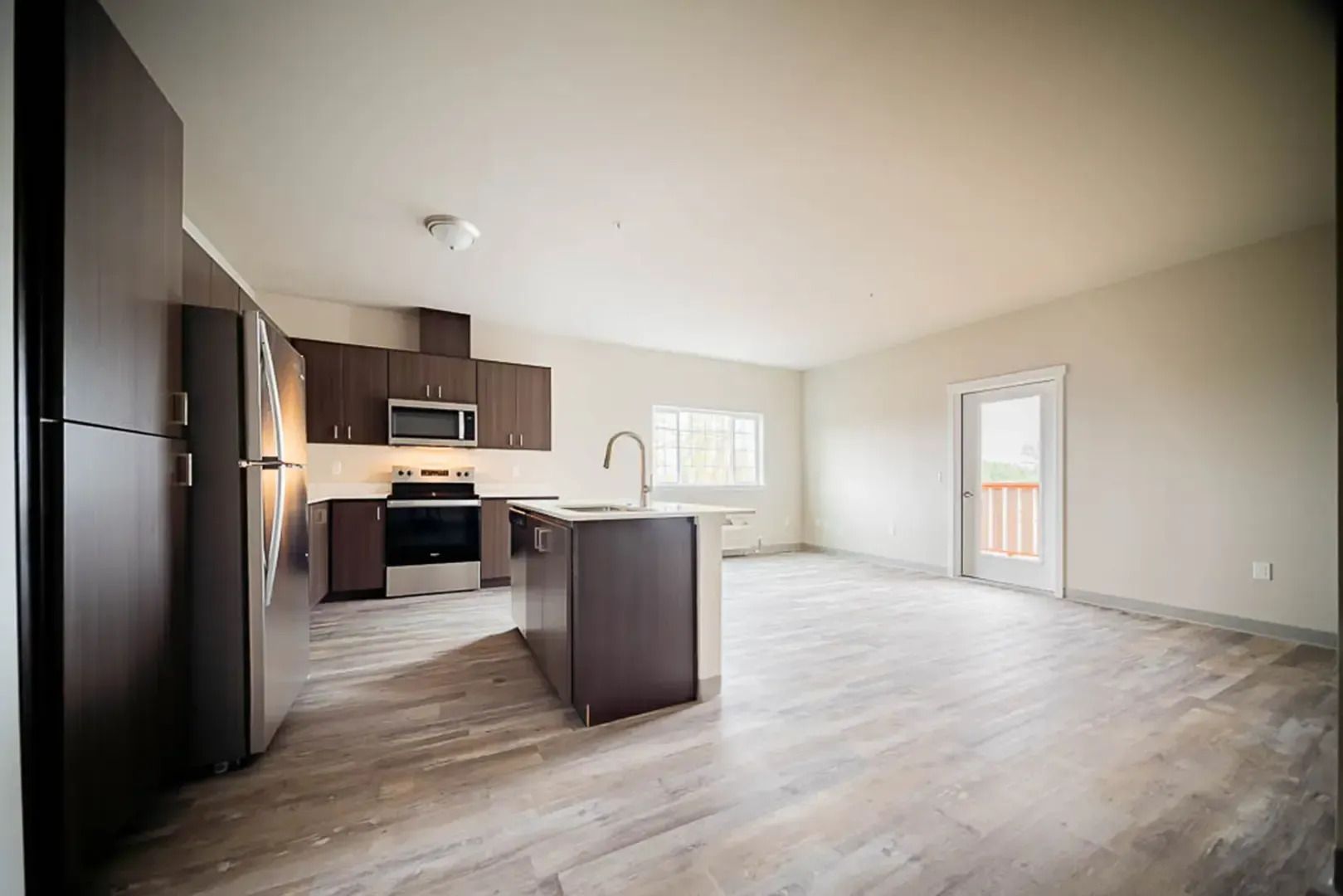 Empty open-plan kitchen and living room with dark cabinets, island, light flooring, and sliding door to balcony