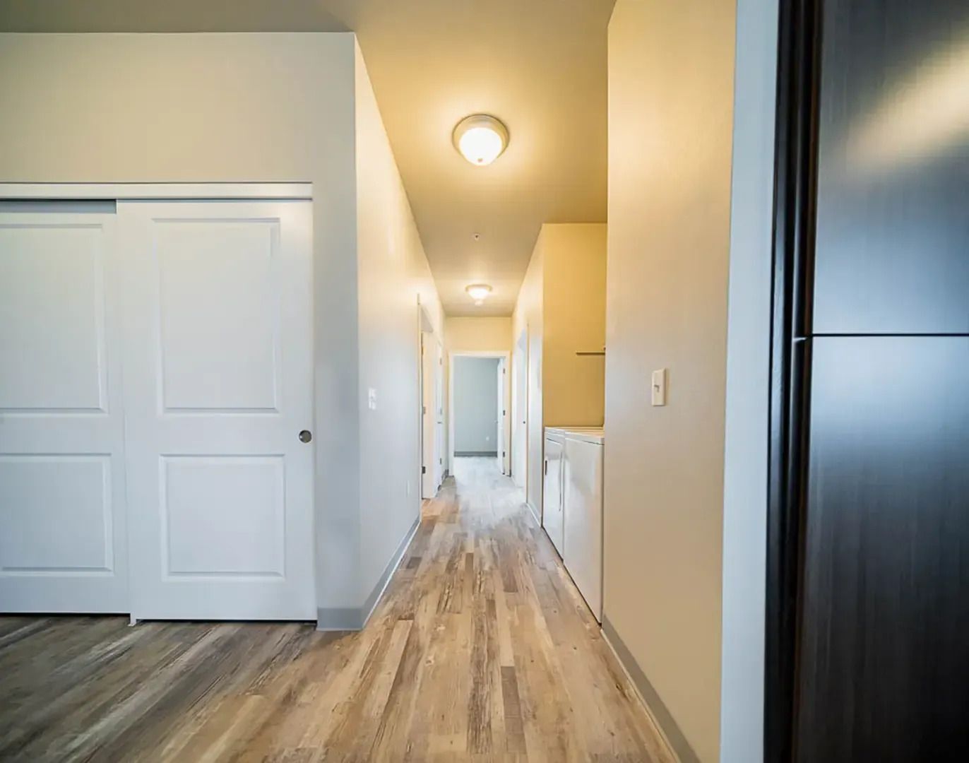 Bright hallway with wood floors, white walls, closet doors, and a kitchen area in the background