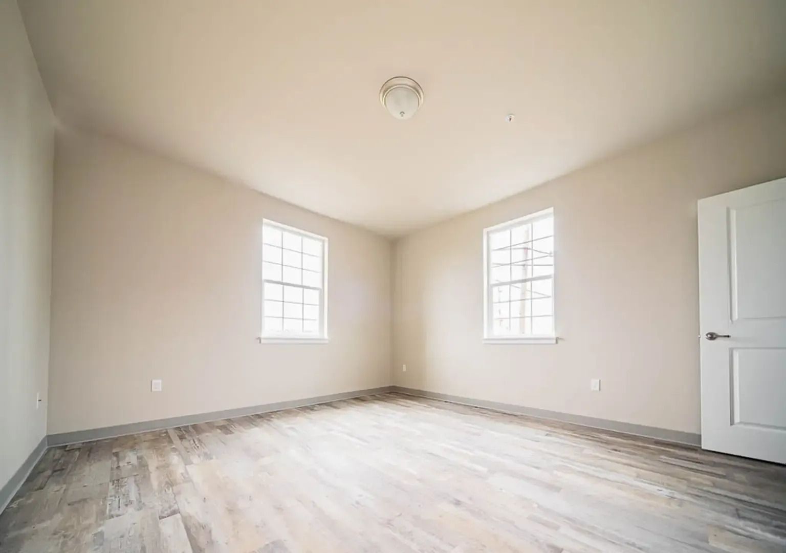 Empty beige bedroom with two windows, white door, and light wood flooring