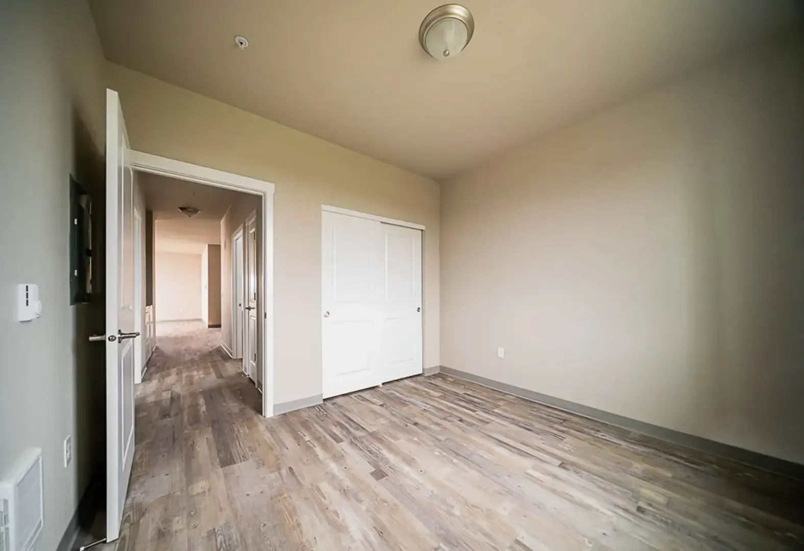 Empty beige bedroom with wood-look flooring, white closet doors, and an open doorway to a hallway