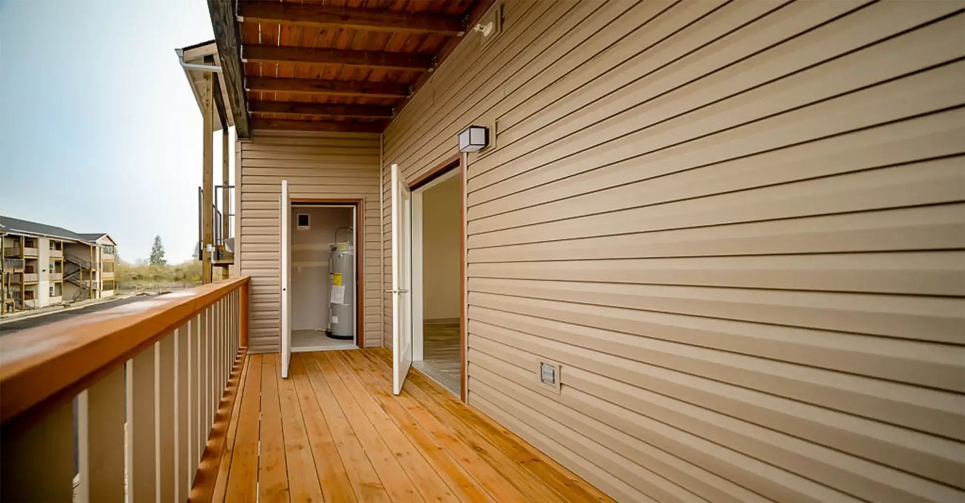 Long wooden balcony with beige siding, railing, and a doorway overlooking nearby apartments