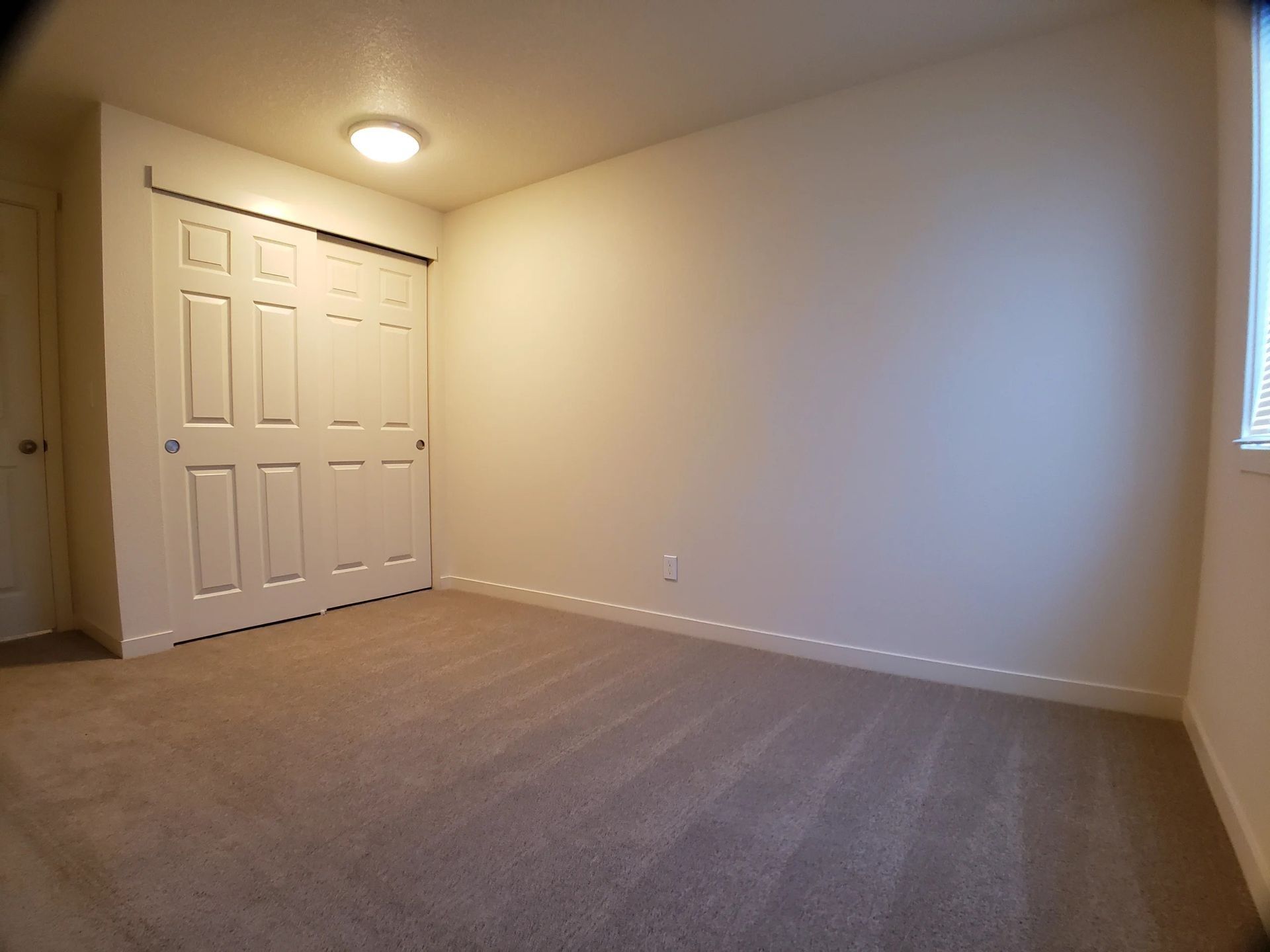Empty beige bedroom with carpet, closed closet doors, and a ceiling light on.