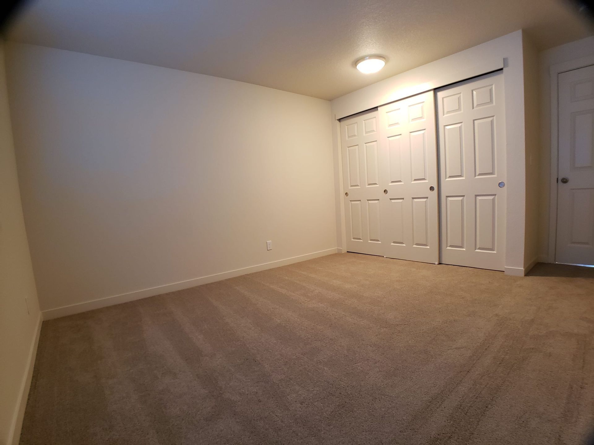 Empty beige bedroom with carpet, white closet doors, and a ceiling light.