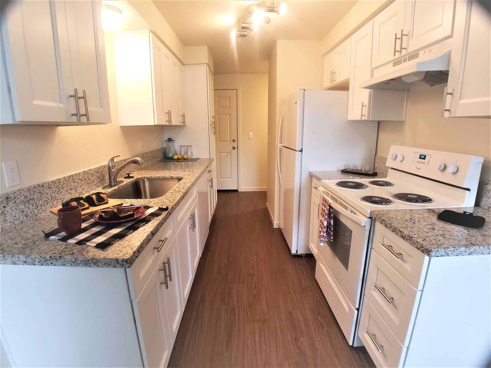 Narrow white kitchen with granite counters, stove, refrigerator, and a long brown wood floor aisle
