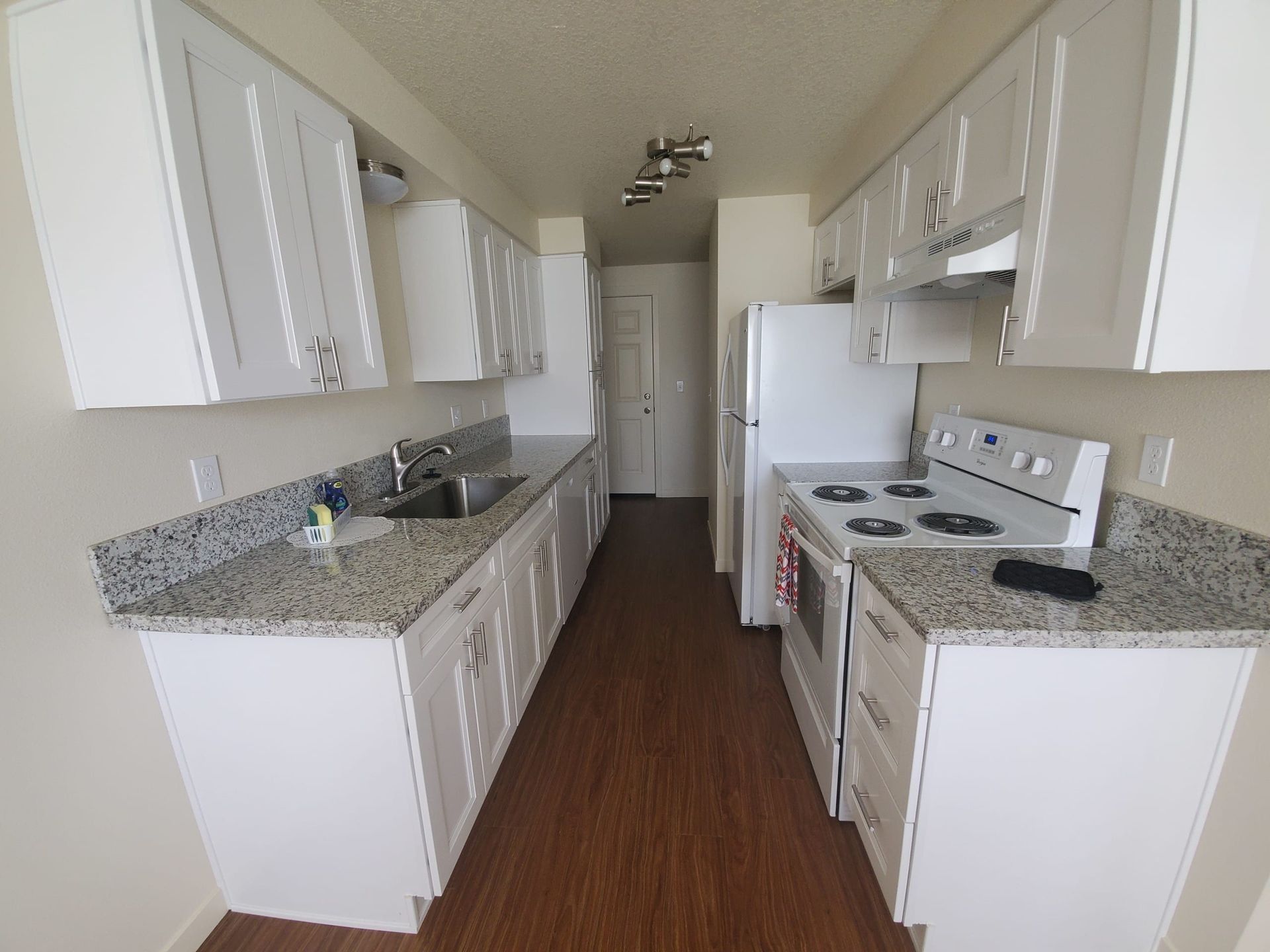 Narrow white kitchen with granite countertops, white cabinets, stove, and refrigerator.
