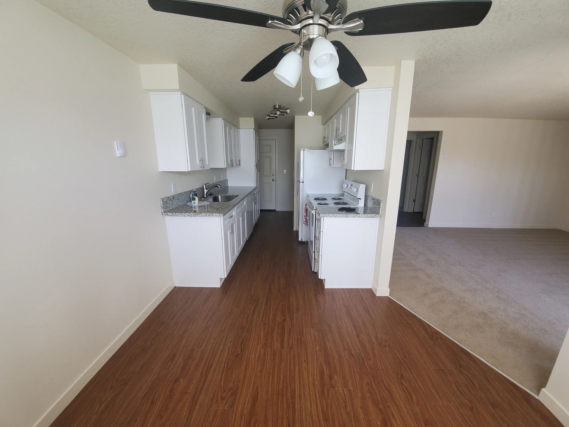Open kitchen with white cabinets, wood floors, ceiling fan, and adjacent carpeted living area.